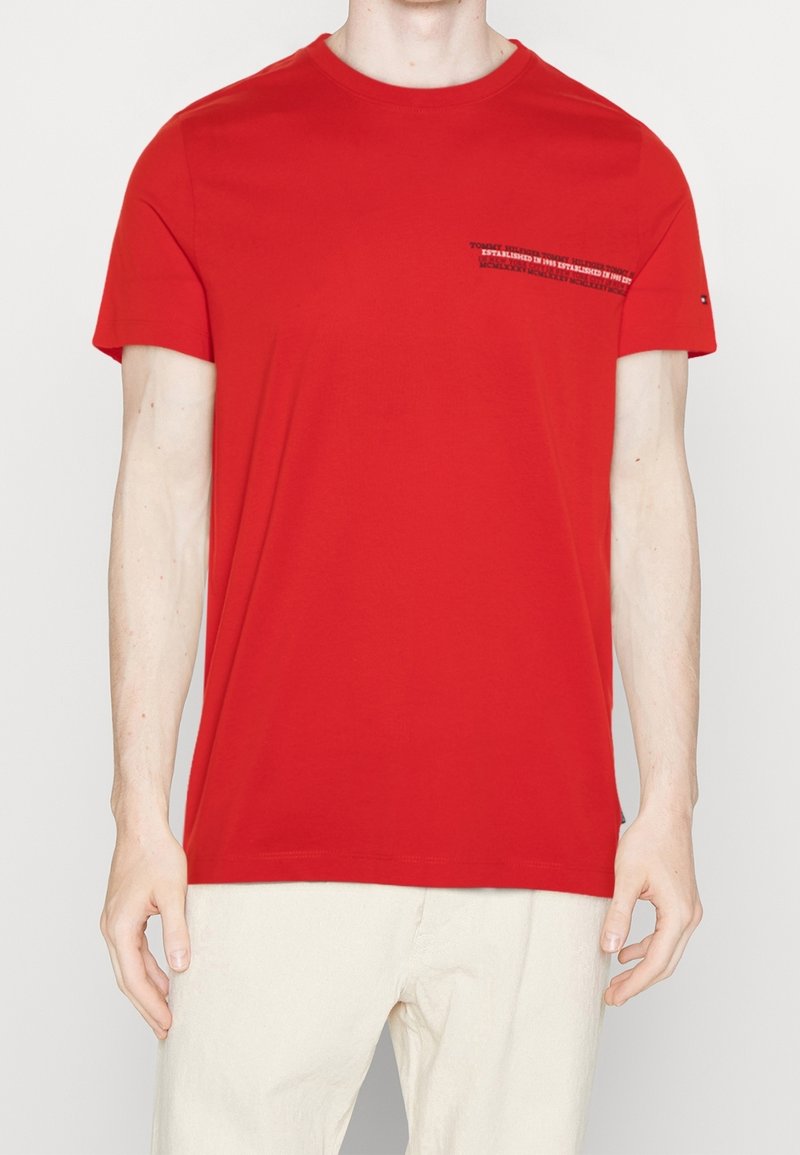 T-shirt en coton rouge avec un col rond et des manches courtes. Présente un texte graphique noir sur la zone de la poche poitrine. Texture lisse.