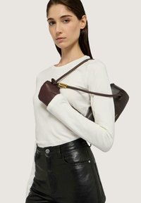 Bolso de cuero marrón con forma redondeada y herrajes dorados, sostenido por una persona que lleva una camisa blanca de manga larga y pantalones de cuero negros.