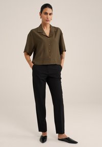 Camicia verde oliva a maniche corte con colletto e chiusura a bottone, abbinata a pantaloni neri su misura. Tessuto strutturato e vestibilità comoda.