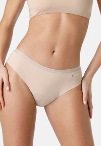 Donna che indossa mutande hipster senza cuciture beige con il logo "LOVABLE" sull'anca su sfondo bianco.