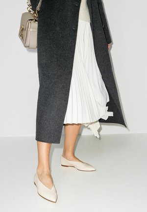 Donna che indossa ballerine beige con punta, gonna midi plissettata bianca, cappotto lungo grigio scuro, con una borsa beige al braccio.