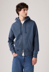 Ung mann med blå zip-up hettegenser med frontlommer og blå jeans, ser til siden mot en ensfarget bakgrunn.