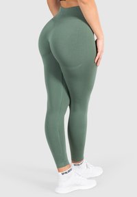 Leggings verdi a vita alta realizzati in tessuto liscio, con una vestibilità aderente e dettagli cuciti, abbinati a scarpe da ginnastica bianche.