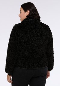 Donna con capelli scuri che indossa una giacca in pile nera a trama e jeans neri, in piedi, con la schiena rivolta verso l'osservatore e la testa girata di lato.