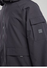 Black jacket featuring a zip front, textured fabric, two front pockets, and a patch on the sleeve with a geometric design.