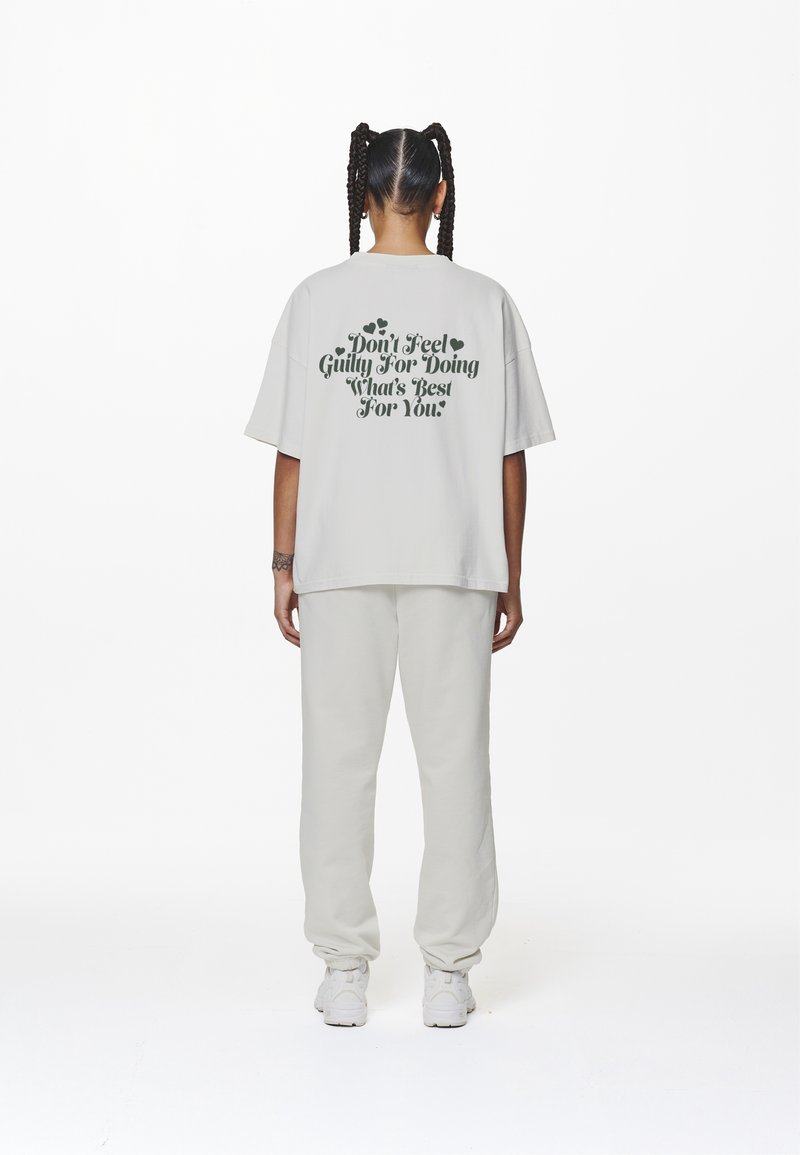 Lös passform ljusgrå t-shirt med mörkgrön text på baksidan som säger "Känn dig inte skyldig för att göra det som är bäst för dig." Enkla vita joggers.