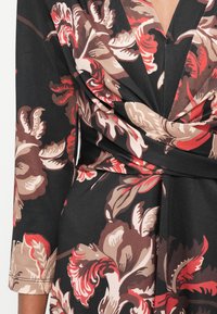 Bloemrijke wikkeljurk met grote rode, beige en bruine bloemen op een zwarte achtergrond. Gemaakt van een textuurstof met een diepe V-hals en lange mouwen.