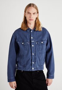 Jeansjacka i mörkblått, med framfickor, knäppning med knappar och långa ärmar. Slät textur med kontrasterande vita knappar.