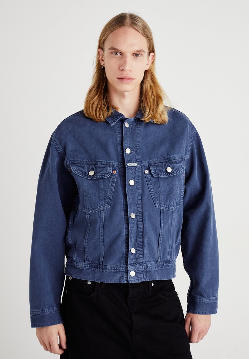 Chaqueta de mezclilla en azul oscuro, con bolsillos frontales, cierre de botones y mangas largas. Textura suave con botones blancos en contraste.
