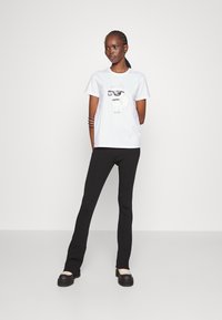 T-shirt alb din bumbac, cu un câine de desene animate purtând ochelari de soare, asortat cu pantaloni negri cu nervuri și pantofi negri masivi.