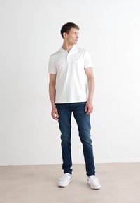 Witte polo met korte mouwen, van katoen, met knoopsluiting bij de kraag, gedragen met blauwe spijkerbroek en witte sneakers. Staand tegen een lichte achtergrond.