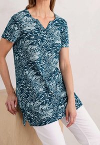 Mujer vistiendo una túnica azul con patrones botánicos y de aves en blanco, combinada con pantalones blancos, sentada sobre un bloque de madera clara.