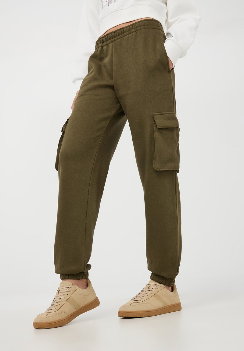Pantaloni felpa verde oliva con polsini affusolati, vita elastica e tasche laterali cargo. Abbinati a sneakers beige per il massimo comfort.