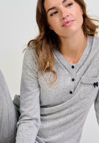 Mujer con una camiseta de manga larga gris claro, con botones negros y un pequeño lazo negro en el bolsillo del pecho, mirando ligeramente de lado.