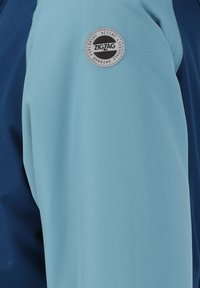 Veste bleue et bleu clair avec un extérieur texturé, un patch logo rond portant l'inscription "ZIGZAG", et des panneaux de couleur contrastée.