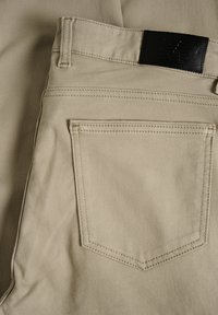 Pantalons beiges avec une texture lisse, comportant une pièce en cuir noire, deux poches arrière et des coutures renforcées le long des coutures.