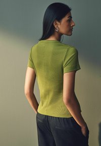 Oliivivihreä ribattua lyhythihaista paitaa, jossa on pyöreä kaula-aukko; teksturoitu kangas, slim fit -malli ja näkyvät saumalähtökohtia takana.
