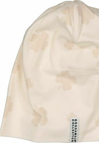 Beige bomullsmössa med subtilt ljusblommönster, rundad topp och en liten etikett i basen som visar "Geggamoja Collection."