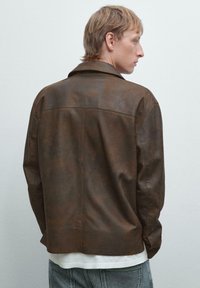 Giacca di pelle marrone con una texture liscia, design con colletto, dettagli delle cuciture visibili e un orlo leggermente curvo, indossata sopra una camicia bianca.