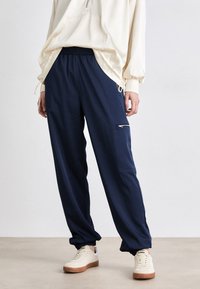 Mørkeblå joggebukser med elastisk midje, sidelommer med glidelås og innsnørte cuffs. Laget av lett, glatt materiale.