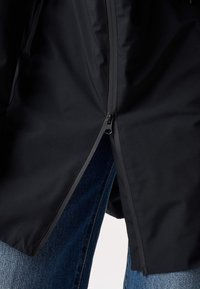 Čierna bunda s čiastočne rozopnutým stredovým zipsom, pod ktorým sú viditeľné modré riflové džínsy.