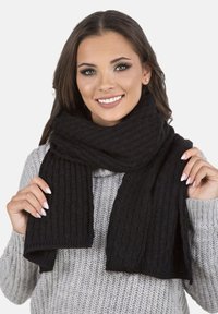 Écharpe tricotée noire avec un motif côtelé texturé, drapée autour du cou, tenue par un mannequin portant un pull gris.