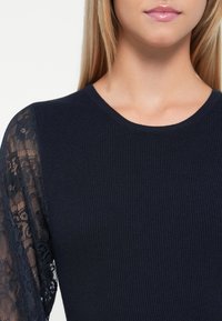 Moteris vilki tamsiai mėlyną megztinį su permatomomis nėriniuotomis rankovėmis, ant kurių pavaizduoti gėlių motyvai, rodoma nuo apykaklės iki vidurinės liemens.