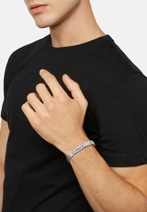 Bracciale d'argento con un design moderno caratterizzato da anelli e un accento con logo, indossato su una mano appoggiata contro una t-shirt nera.