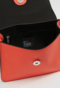 Sac à main en cuir rouge au toucher lisse, avec un fermoir à pression en argent et une poche zippée intérieure noire. Étiquette de logo visible à l'intérieur.