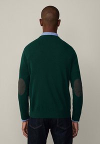 Vihreä villapaita, jossa on pyöreät mustat kyynärpäälaput, pyöreä pääntie ja pitkät hihat, kerroksittain vaaleansinisen paidan päällä, yhdistettynä tummiin farkkuihin.