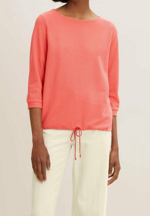 Mujer con top coral de manga larga de punto con cordón en el dobladillo y pantalones blancos, de pie contra un fondo claro y liso.