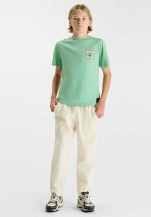 Adolescent portant un t-shirt vert à manches courtes avec un écusson, un pantalon couleur crème et des baskets noir et blanc, debout les mains dans les poches.