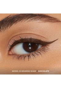 Close-up van een oog met een donkerbruine iris en wimpers. Oogschaduw in een chocolade tint creëert een scherpe vleugelvorm. De huid heeft een soepele textuur.