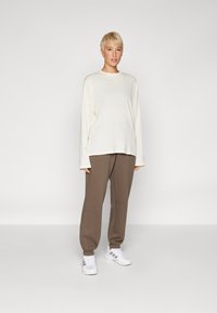 Langermet kremfarget topp, avslappet passform, kombinert med brune smale joggebukser med elastiske mansjetter og hvite treningssko. Enkelt design, myk tekstur.
