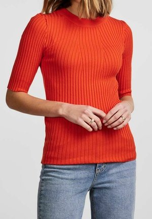Femme portant un haut ajusté à manches courtes en côtes, de couleur orange vif, associé à un jean bleu clair, montrant des bagues aux deux mains.