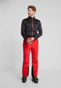 Röda skidbyxor med svarta sidopaneler, kombinerat med en svart zip-up-jacka med röda detaljer och dragkedja. Jackan har två framfickor.
