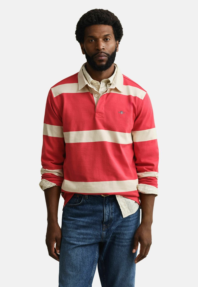 Mann mit Bart, der ein rot-beige gestreiftes Rugbyhemd über einem beigen Hemd mit Kragen und blauen Jeans trägt, vor einem weißen Hintergrund stehend.