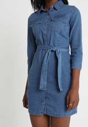 Kobieta w niebieskiej sukience dżinsowej zapinanej na guziki, z długimi rękawami, przednimi kieszeniami i wiązanym paskiem w talii, na jednolitym tle.