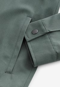 Abrigo de tela verde con una textura suave, que presenta un puño con un botón a presión negro y costuras limpias a lo largo de los bordes.