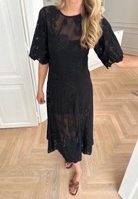 Mustat midi-mekko, jossa on läpinäkyvät kukkakuvioiset hihat ja puku, vyötärö on istuva ja hame on laskostettu, yhdistettynä ruskeisiin liukusandaaleihin.