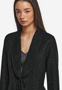 Svart texturerad kofta med sjalkrage och knytning, med intrikata mönster och subtil glans genom hela tyget.