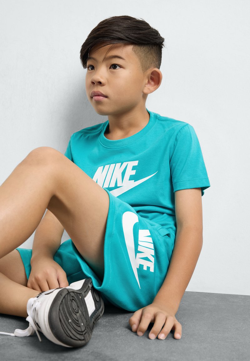 Tyrkysový set trička a šortek Nike, s velkými bílými potisky loga. V sedící pozici, s černými botami a texturovanou šedou podlahou.