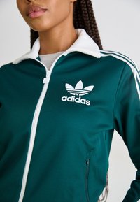 Casaco verde com fecho zip, colarinho e riscas brancas; apresenta o logo da Adidas e uma textura suave; inclui bolsos laterais.