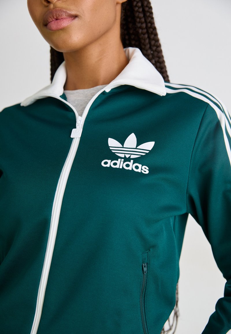 Grüne Zip-Jacke mit weißem Kragen und Streifen; verfügt über das Adidas-Logo und eine glatte Textur; beinhaltet seitliche Taschen.