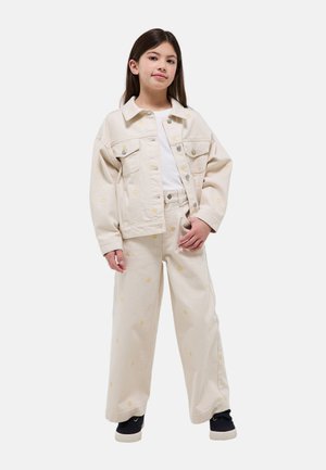 Fille debout portant une veste beige et un pantalon large imprimé de petites fleurs jaunes, chemise blanche et baskets noires.