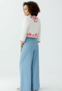 Witte cropped jas met rode bloemenborduring en bies, gecombineerd met lichtblauwe hoge taille flared jeans. Soepele textuur, casual ontwerp.