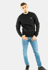 Sudadera negra con cuello redondo y detalles de rayas blancas en los puños, combinada con jeans de mezclilla azul claro y zapatos negros. Presenta un logo en el pecho.