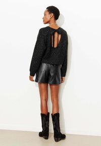 Pull court noir scintillant avec une ouverture au dos nouée par un ruban, associé à un short en cuir noir et des bottes noires montantes ornées.