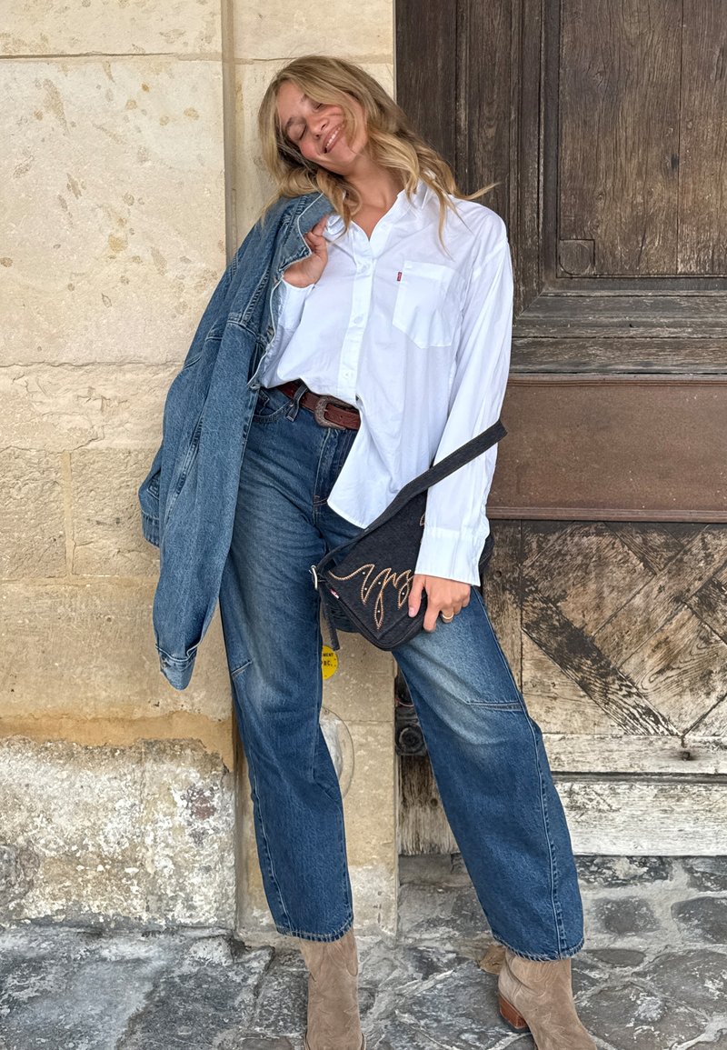 Camisa branca de botões, jaqueta de ganga azul, jeans largos azuis, botas de tornozelo castanhas e uma bolsa preta de mensageiro com costura decorativa.