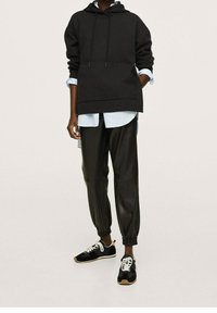 Sudadera negra de gran tamaño con bolsillo canguro, superpuesta a una camisa de botones azul claro. Joggers de cuero negros y zapatillas negras con cordones blancos.